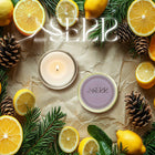 SerrBeauty Cedarwood, Fir & Lemon Beeswax Scented Candle 125 G in a round tin, surrounded by fresh lemons, fir branches, and pinecones. A natural, organic candle with a refreshing woodsy and citrus aroma.