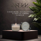 SerrBeauty Eucalyptus, Cinnamon, Clove & Lemon Beeswax Scented Candle in a glass jar with a lit wick, placed on a dark platform. Organic and handcrafted for a refreshing and warm home ambiance.