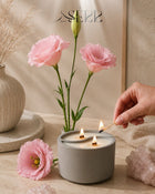 Gray concrete candle with wood wicks, surrounded by pink flowers, cozy home ambiance