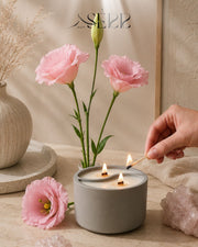 Gray concrete candle with wood wicks, surrounded by pink flowers, cozy home ambiance